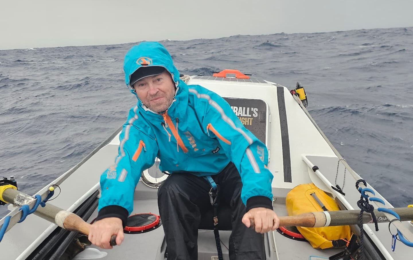 An ocean rower in wet weather clothing rowing at sea.