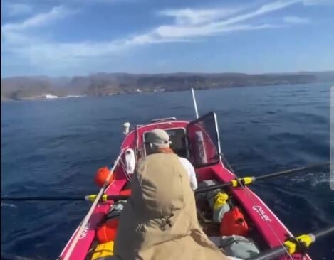 From the bow cabin of an ocean rowing boat, looking back at the 2 rowers on the oars and land in the distance